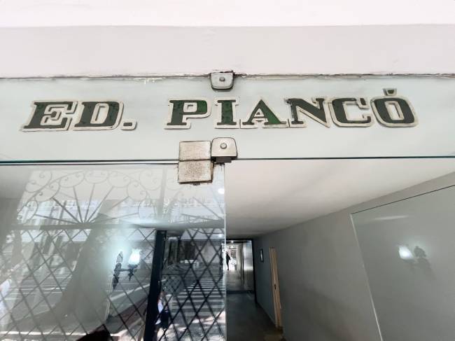 Placa de vidro com ED. PIANC&Oacute; em letras verdes escuras e borda branca, acima de uma porta de vidro com grades e reflexos da rua, e um corredor cinza &agrave; direita