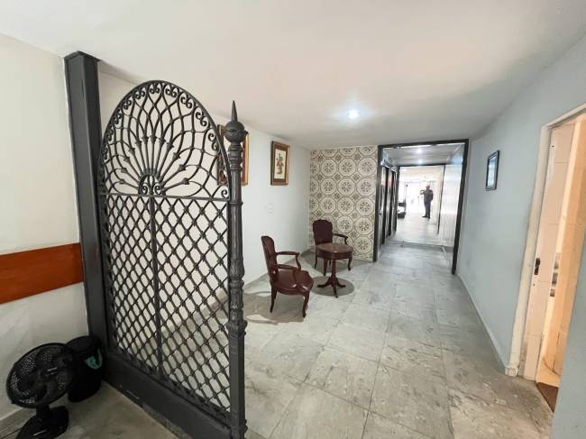 Corredor com port&atilde;o de ferro forjado &agrave; esquerda, ventilador no ch&atilde;o, duas poltronas e uma mesa de madeira escura, parede com azulejos decorados e outra lisa, e uma pessoa ao fundo de um corredor espelhado
