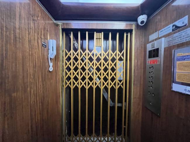 Interior de um elevador antigo com paredes de madeira escura e porta sanfonada dourada. &Agrave; esquerda, um interfone branco. &Agrave; direita, um painel de bot&otilde;es prateado e avisos fixados. Uma c&acirc;mera de seguran&ccedil;a branca est&aacute; no canto superior direito