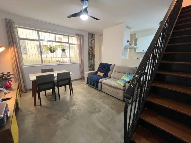 Sala de estar e jantar integradas com sof&aacute; bege, manta azul, almofadas coloridas, mesa de jantar com quatro cadeiras escuras, e escada de madeira com corrim&atilde;o preto &agrave; direita. Uma janela grande com grades e cortinas claras, e um ventilador de teto preto iluminado, completam o ambiente. Ao fundo, uma cozinha clara &eacute; vis&iacute;vel