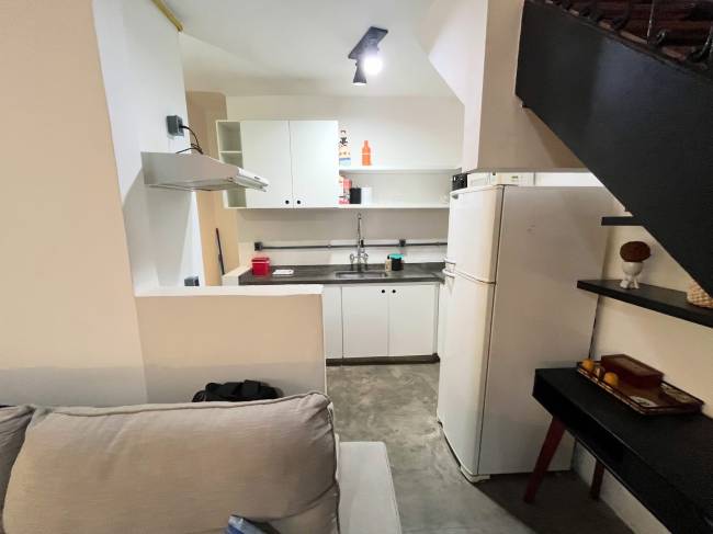 Cozinha compacta com arm&aacute;rios brancos, bancada escura com pia e fog&atilde;o, geladeira branca e exaustor met&aacute;lico. &Agrave; direita, uma escada preta e uma mesa de cabeceira escura com objetos decorativos.