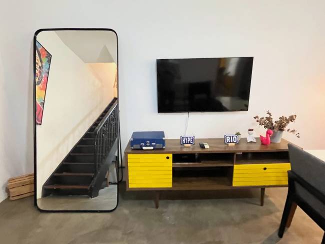 Espelho de corpo inteiro com moldura preta refletindo uma escada escura e um quadro colorido. &Agrave; direita, uma televis&atilde;o preta na parede acima de um rack de madeira com portas amarelas. Sobre o rack, um toca-discos azul, placas HYPE e RIO, e um vaso com plantas secas e um flamingo rosa decorativo. O ch&atilde;o &eacute; de cimento queimado e h&aacute; caixas de madeira empilhadas no canto esquerdo