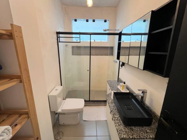 Banheiro moderno com paredes claras, chuveiro de box com estrutura preta, vaso sanit&aacute;rio branco, bancada de granito cinza e pia preta retangular. Acima da pia, um arm&aacute;rio espelhado com prateleiras pretas. &Agrave; esquerda, uma estante de madeira clara com prateleiras vazias. Uma janela retangular alta com vista para o c&eacute;u azul est&aacute; acima do box. Uma l&acirc;mpada pendente ilumina o ambiente