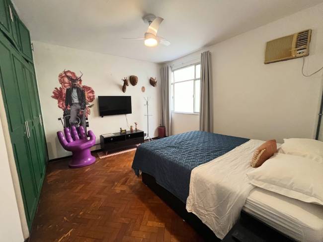 Quarto com cama de casal coberta por edredom azul e len&ccedil;&oacute;is brancos, arm&aacute;rio verde, poltrona roxa em formato de m&atilde;o, e adesivo de parede com homem de cabe&ccedil;a de veado e flores. H&aacute; uma TV preta, um rack, um cabideiro e um ar-condicionado na parede branca, al&eacute;m de um ventilador de teto e piso de madeira