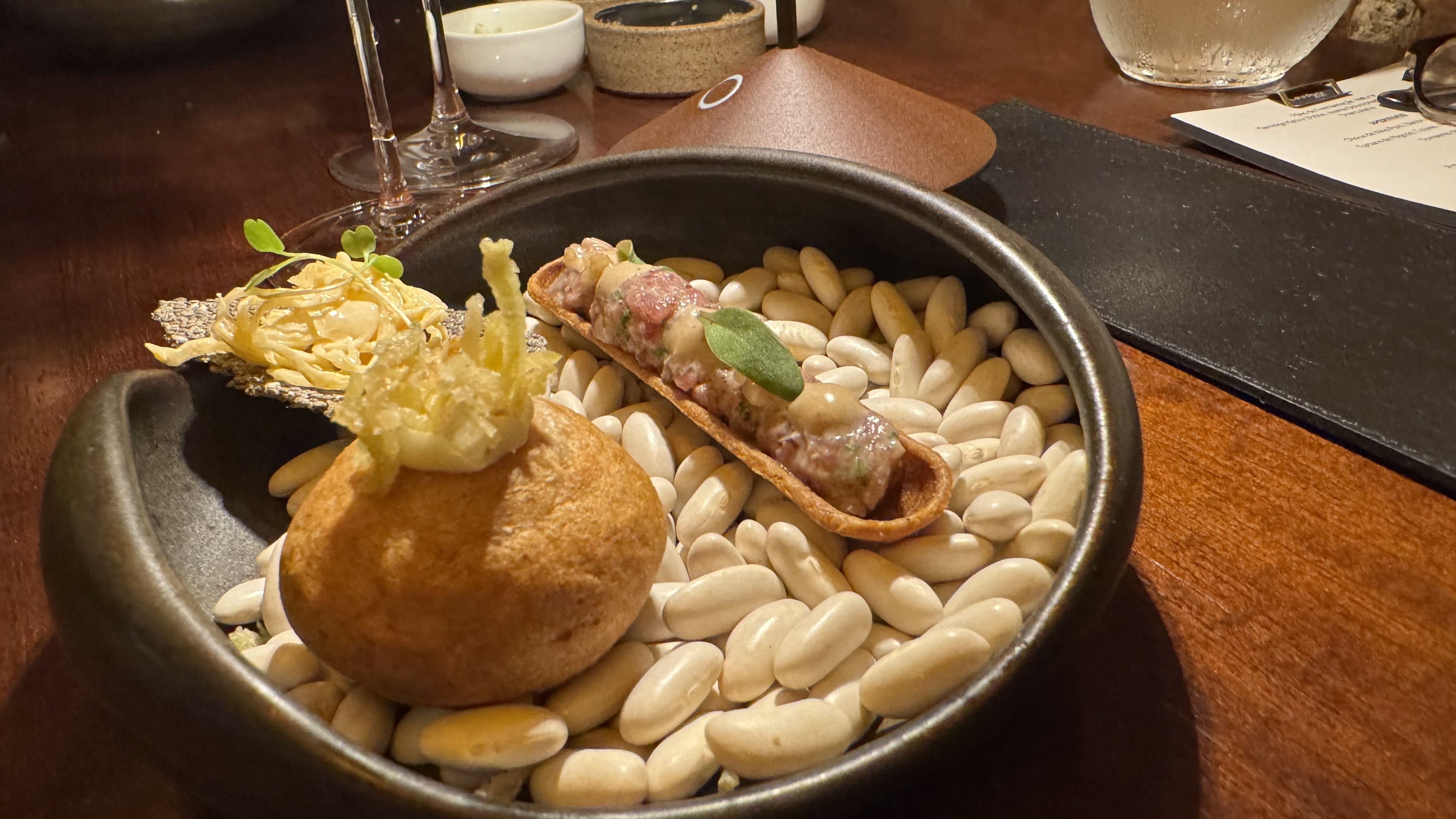 Prato gourmet em tigela escura com feijões brancos, um bolinho dourado com crocante, um taco com recheio de carne e folha verde, e massa fina ao lado. O cenário é uma mesa de madeira com taças e um menu
