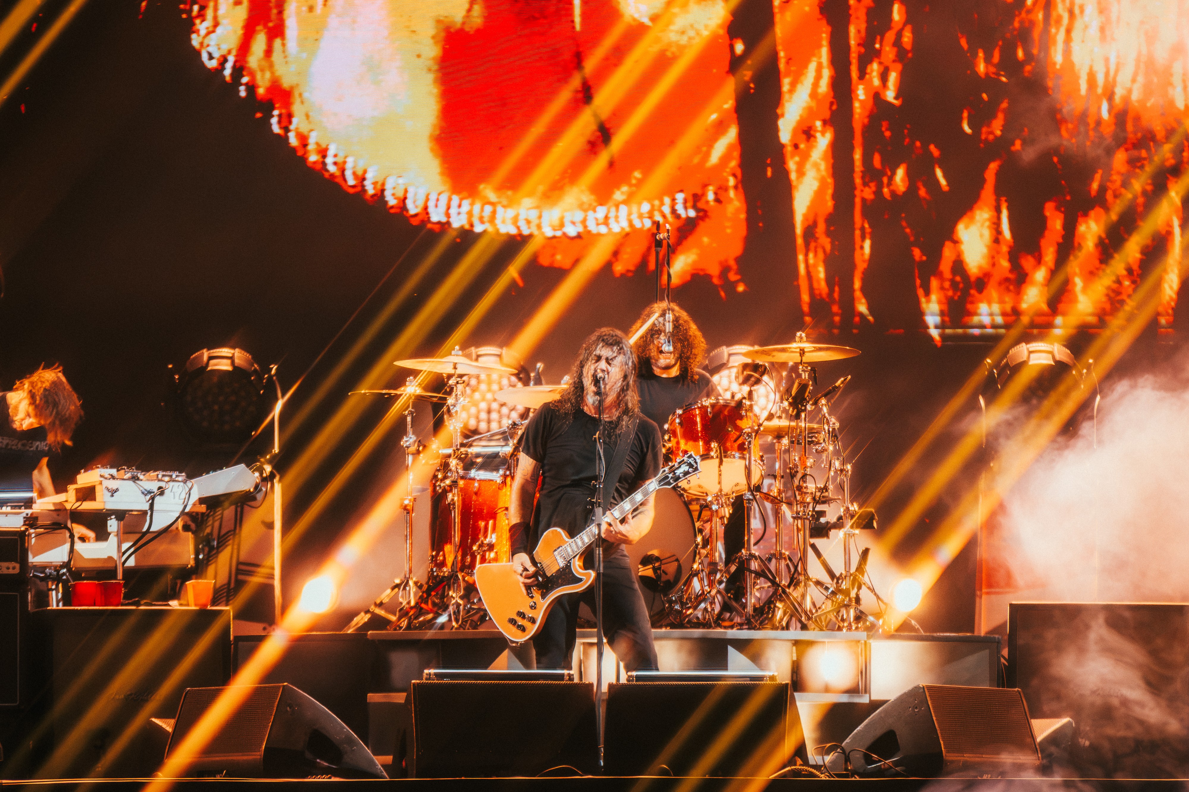Cantor de rock com cabelo comprido e camiseta preta, tocando guitarra e cantando em um palco iluminado por luzes laranja e amarelas, com um baterista ao fundo e uma tela exibindo chamas