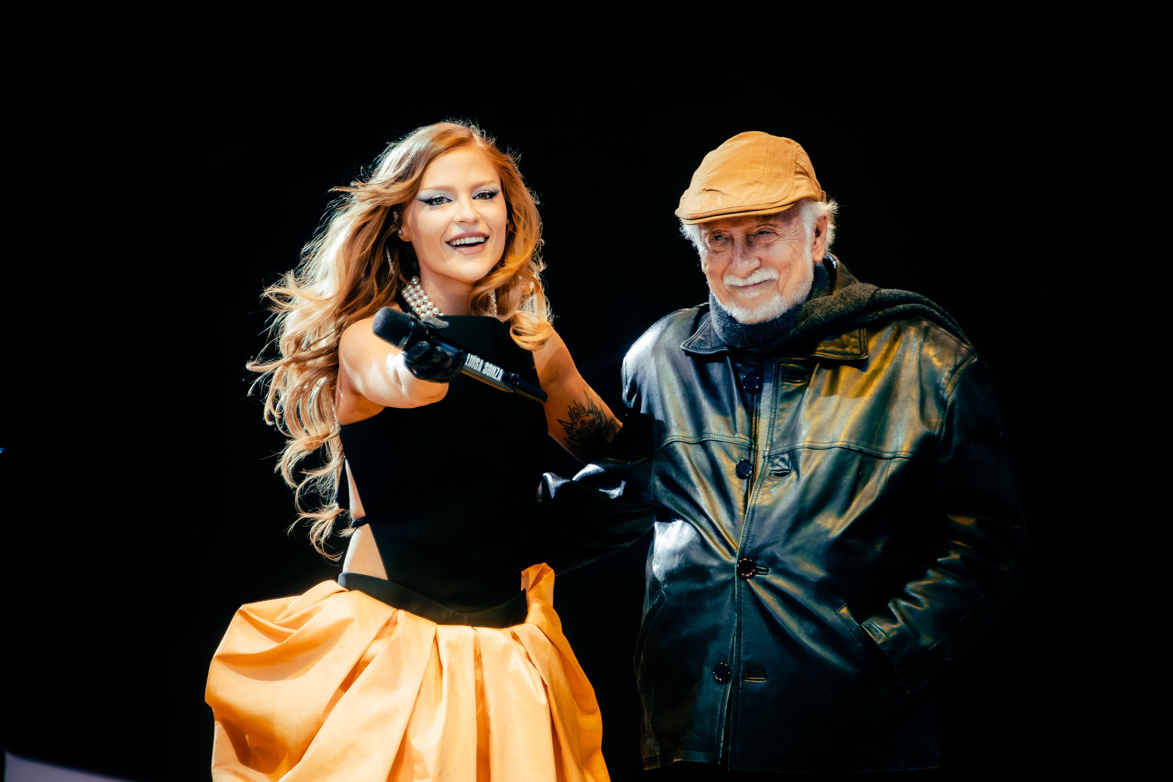 A cantora Marina Sena, sorrindo, com cabelo ruivo solto, blusa preta e saia laranja bufante, aponta um microfone para frente. Ao lado, o cantor e compositor Chico Buarque, sorrindo, usa boina bege, cachecol escuro e jaqueta de couro preta. Ambos estão em um palco escuro