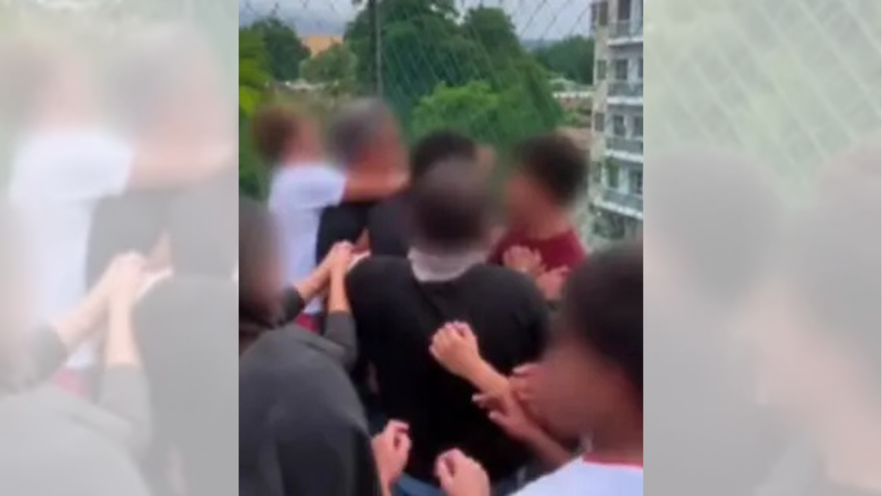 Grupo de jovens com rostos borrados, alguns vestindo camisetas escuras e outros claras, aglomerados em uma briga. Mãos e braços se estendem, indicando confronto físico. Ao fundo, uma cerca de tela, árvores e um prédio de apartamentos, sugerindo um ambiente urbano ou escolar
