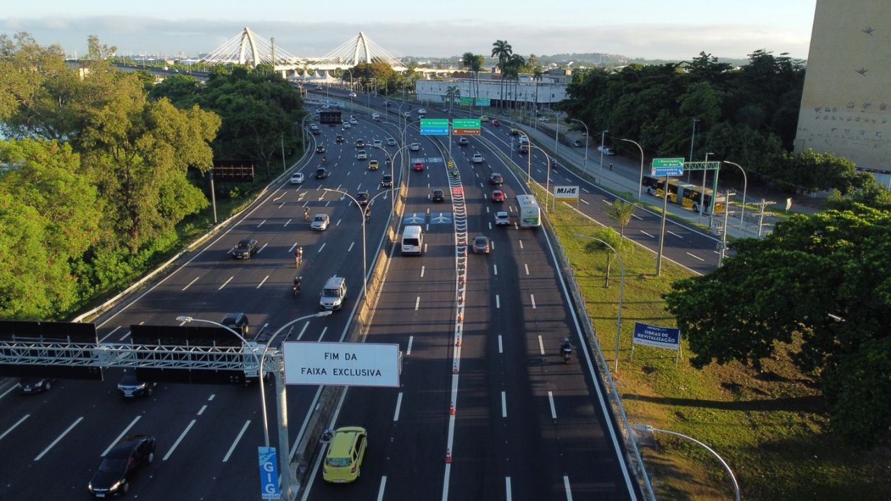 Vista aérea de uma rodovia movimentada com vários carros e motos, sob um céu claro. Há uma placa branca à esquerda indicando FIM DA FAIXA EXCLUSIVA. Ao fundo, uma ponte estaiada e vegetação densa margeiam a pista