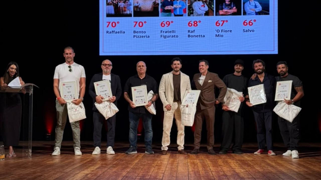 Um grupo de oito homens e uma mulher em um palco de madeira, recebendo prêmios. A mulher, à esquerda, anota em uma prancheta. Os homens seguram certificados e sacolas brancas. Ao fundo, um telão exibe uma lista de nomes e números, como 70° Raffaella e 66° Salvo