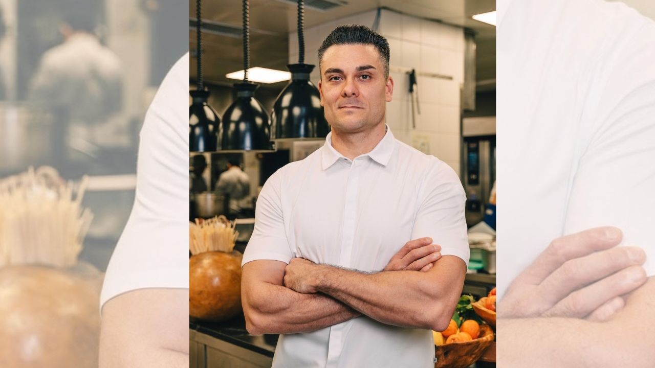 Homem branco de cabelo escuro e camisa branca, braços cruzados, sorri levemente para a câmera em uma cozinha profissional com iluminação pendente e utensílios ao fundo