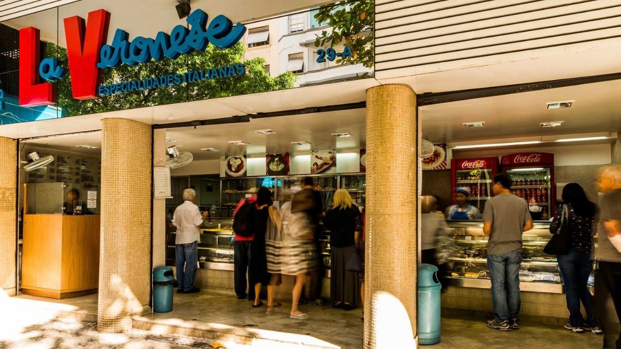 Fachada de uma loja chamada La Veronese Especialidades Italianas, com pessoas em pé dentro do estabelecimento, algumas com movimento borrado, e um balcão de atendimento à esquerda. O letreiro da loja tem letras vermelhas e azuis, e há um número 29-A na parte superior direita. Duas lixeiras azuis estão visíveis, uma em cada lado da entrada.