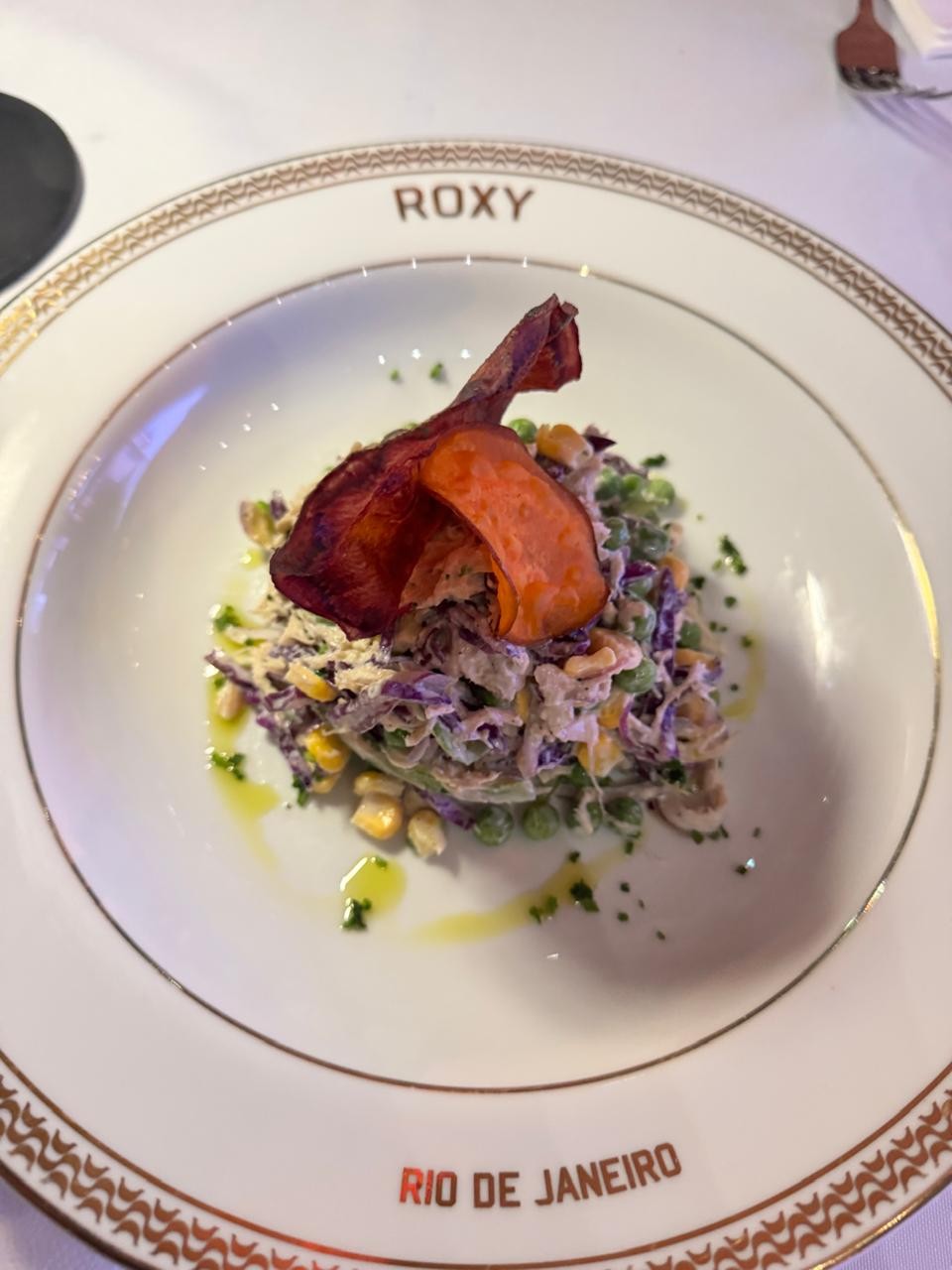 Salada colorida com repolho roxo, milho, ervilhas e molho cremoso, decorada com chips de batata doce e gotas de azeite, servida em prato branco com borda dourada e os dizeres ROXY e RIO DE JANEIRO