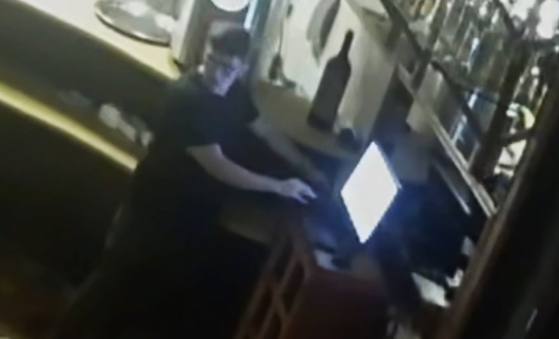 Homem de óculos e camiseta escura, inclinado sobre um balcão, manipulando um objeto brilhante à direita, em ambiente interno com garrafa e prateleiras ao fundo