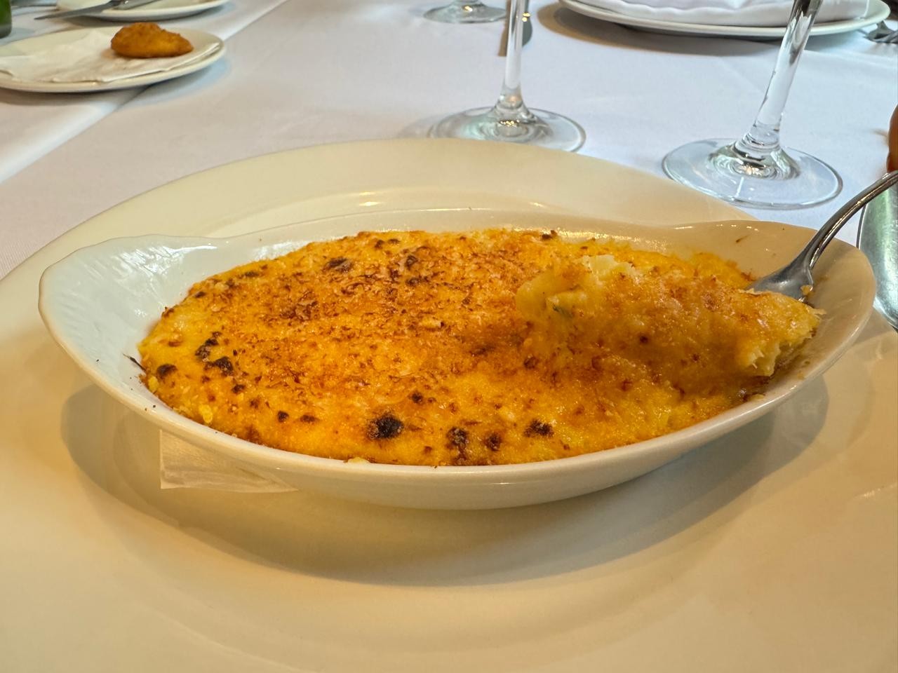 Travessa branca oval com macarrão gratinado, coberto por uma crosta dourada e crocante. Uma colher de metal retira uma porção cremosa do prato, que está sobre um prato branco maior em uma mesa com toalha branca. Ao fundo, copos de vinho e um pequeno salgado.