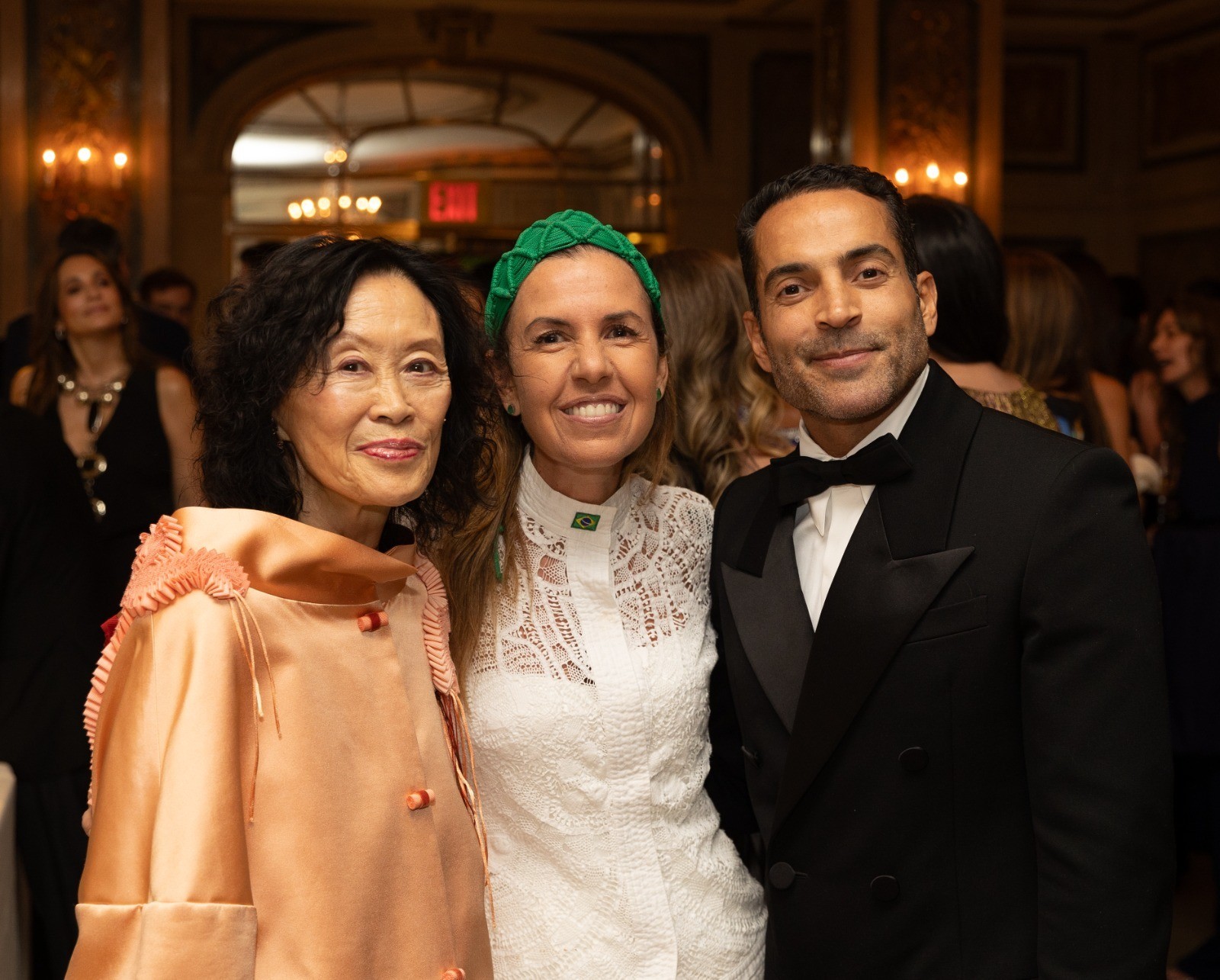 Três pessoas sorrindo para a câmera em um evento formal. Uma mulher asiática de casaco salmão com babados, uma mulher de blusa branca de renda e tiara verde, e um homem de smoking preto e gravata borboleta.