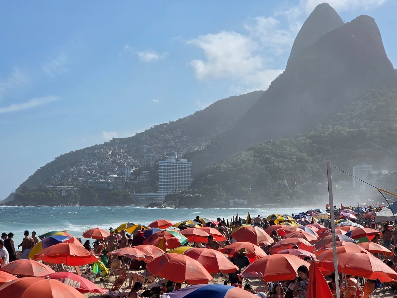 Praia lotada com guarda-sóis coloridos e pessoas, o mar ao fundo, e montanhas com casas e prédios sob um céu azul claro