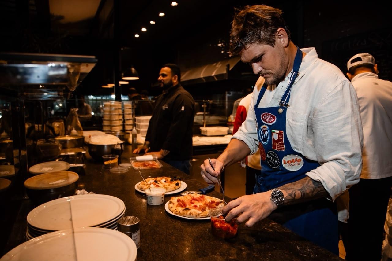 Um chef de camisa clara e avental azul com broches, tatuagem no braço e relógio, prepara pizzas em uma bancada escura. Ele usa um garfo para pegar um ingrediente vermelho de um pote transparente. Ao fundo, outro chef de camisa preta e um terceiro de camisa branca e boné trabalham em uma cozinha com pouca luz e utensílios de metal. Há pratos empilhados e pizzas prontas na bancada