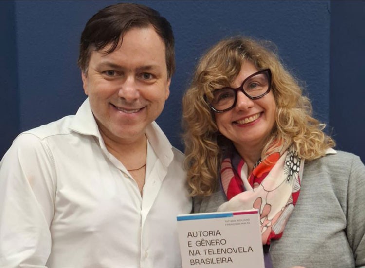 Homem e mulher sorrindo para a câmera, ele de camisa branca e ela de óculos, lenço colorido e blusa cinza, segurando o livro Autoria e Gênero na Telenovela Brasileira