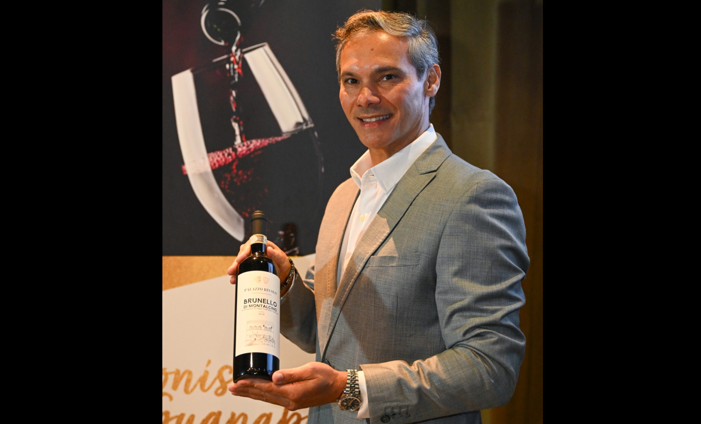 Homem de terno cinza e camisa branca sorri, segurando uma garrafa de vinho tinto Brunello di Montalcino. Ao fundo, um banner com uma taça de vinho e a bebida sendo servida
