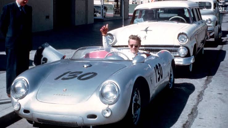 James Dean sorrindo e acenando com luva vermelha dentro de um carro de corrida prateado com o número 130, ao lado de um carro branco e um homem de terno escuro, em uma rua ensolarada