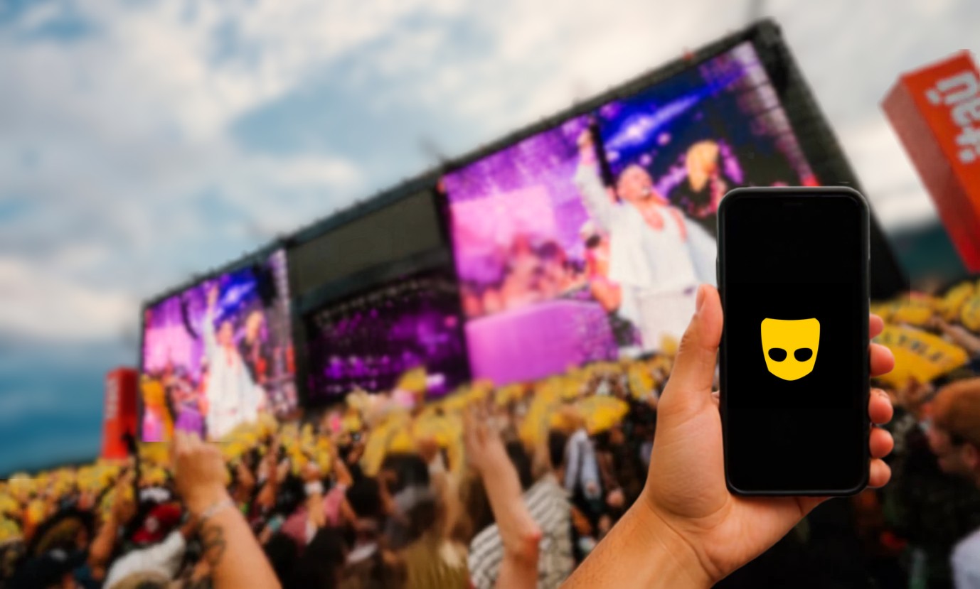 Mão segura celular com o aplicativo Grindr aberto, em primeiro plano. Ao fundo, um show com telões exibindo um cantor e uma multidão com as mãos para cima, em um festival a céu aberto