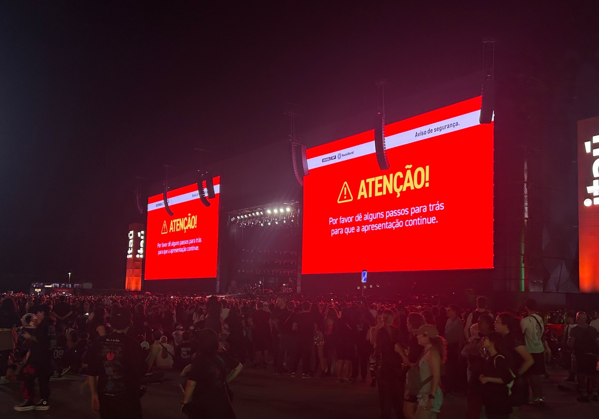 Multidão em show noturno com dois telões vermelhos exibindo ATENÇÃO! Por favor dê alguns passos para trás para que a apresentação continue