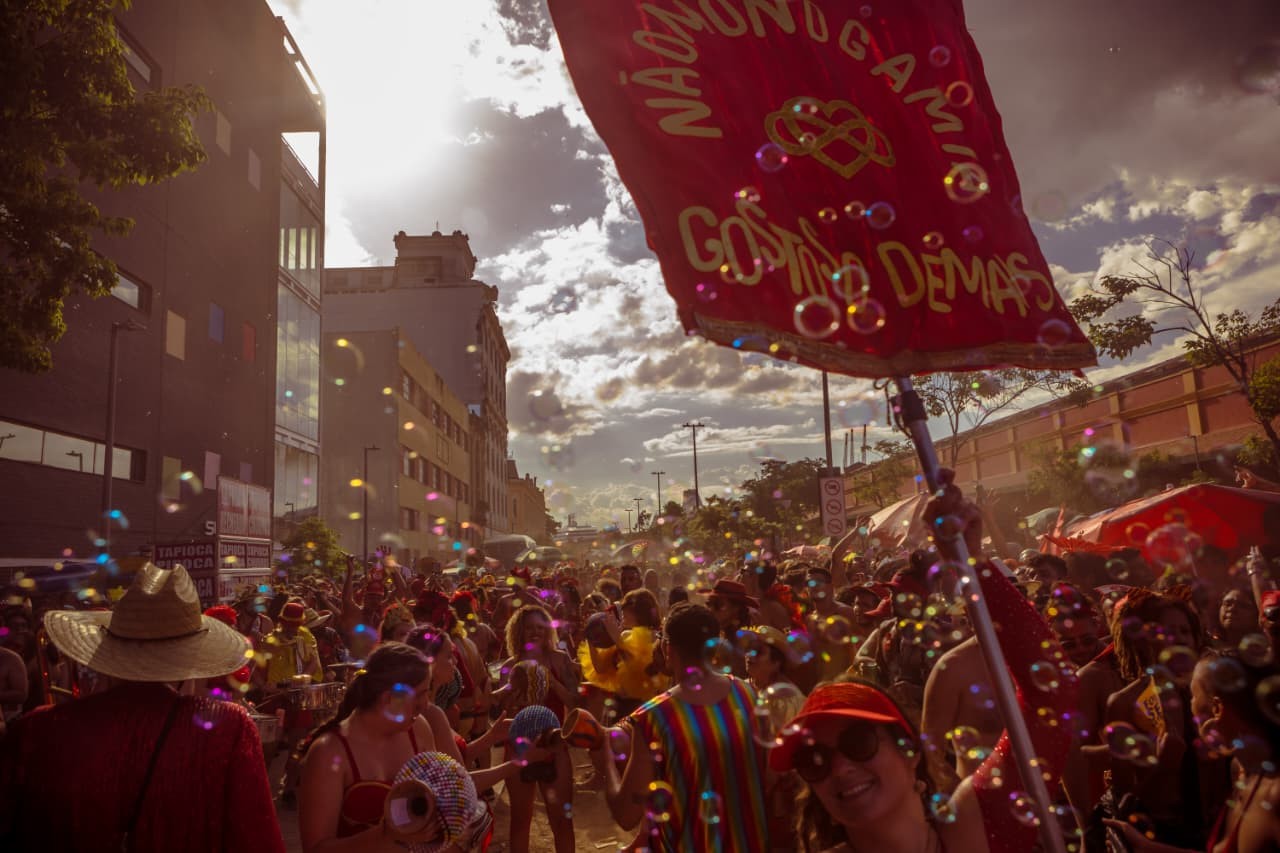 Multidão em festa de rua, com muitas pessoas coloridas e bolhas de sabão flutuando. Um estandarte vermelho com a frase NÃO MUNDANO, GAMIA, GOSTOSO DEMAIS é visível no canto superior direito. O sol brilha forte no céu parcialmente nublado, iluminando edifícios ao fundo. Uma mulher de boné vermelho sorri para a câmera.