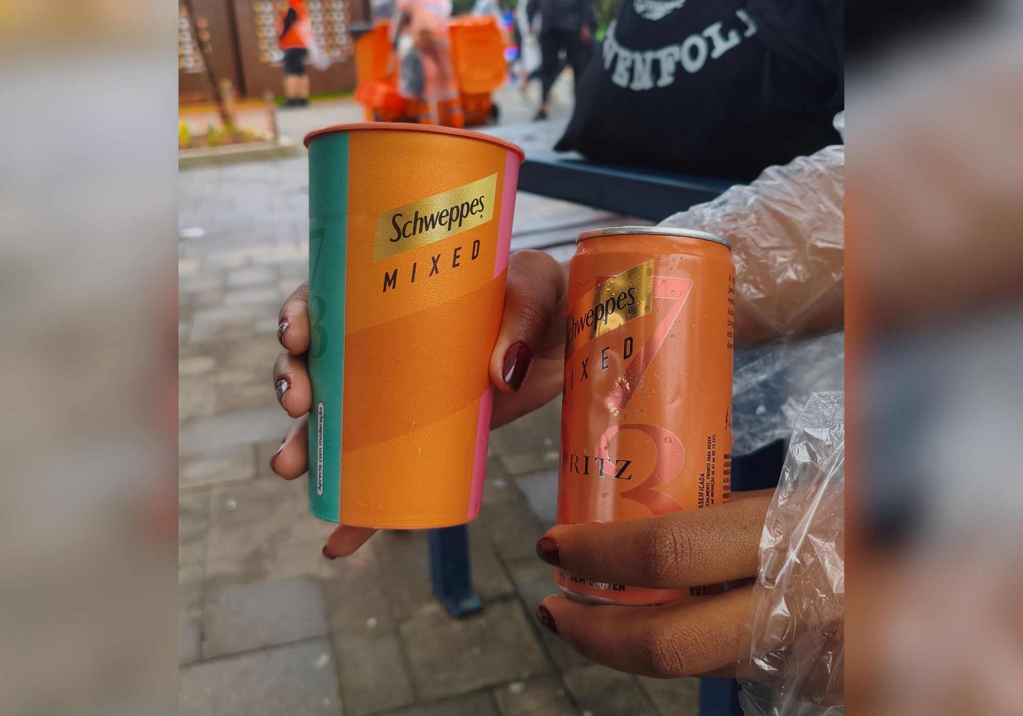 Duas mãos seguram um copo e uma lata de Schweppes Mixed, ambos em tons de laranja, verde-água e rosa. As unhas estão pintadas de vinho. Ao fundo, pessoas e objetos desfocados em um ambiente externo