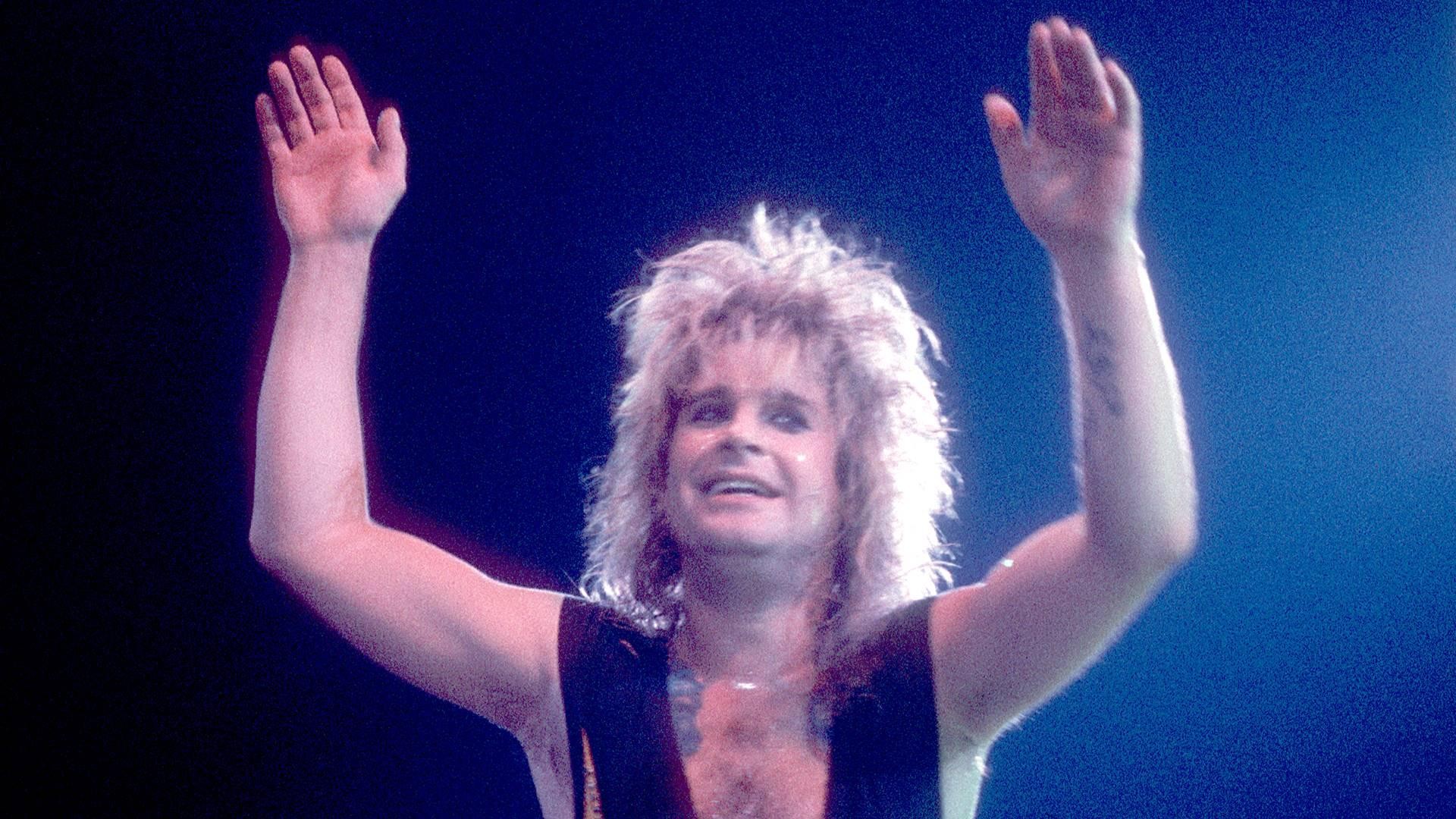 Ozzy Osbourne, com cabelos loiros e volumosos, sorri e levanta os dois braços abertos para cima, em um palco com iluminação azul