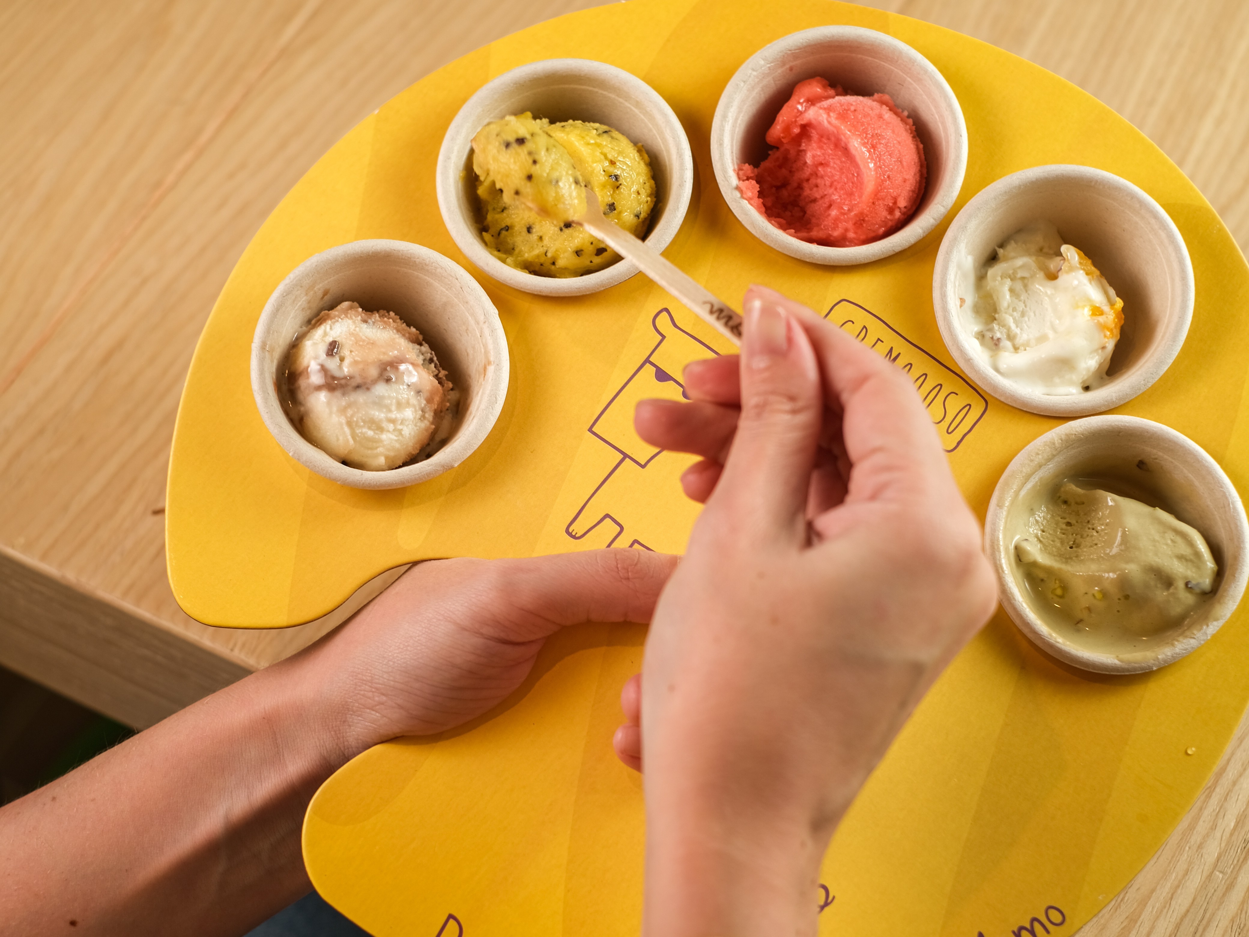 Mãos de uma pessoa segurando uma pequena colher de madeira para provar sorvetes em um suporte amarelo. Há cinco potinhos brancos com sorvetes de diferentes sabores: um amarelo com pontos pretos, um vermelho, um branco com pedaços marrons, um branco com pedaços alaranjados e um verde claro