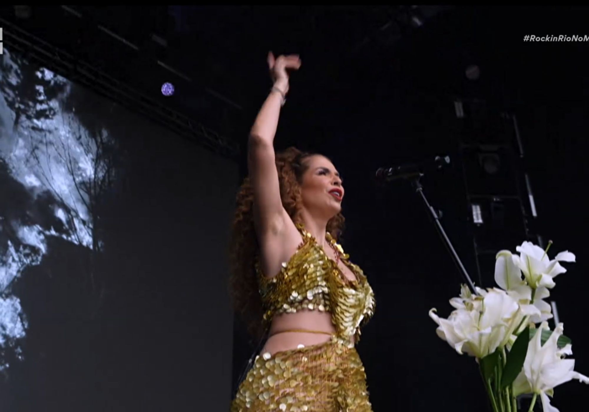 Cantora de cabelos cacheados ruivos, vestindo top e saia dourados de lantejoulas, braço direito erguido, cantando em palco escuro com projeção abstrata e flores brancas à direita