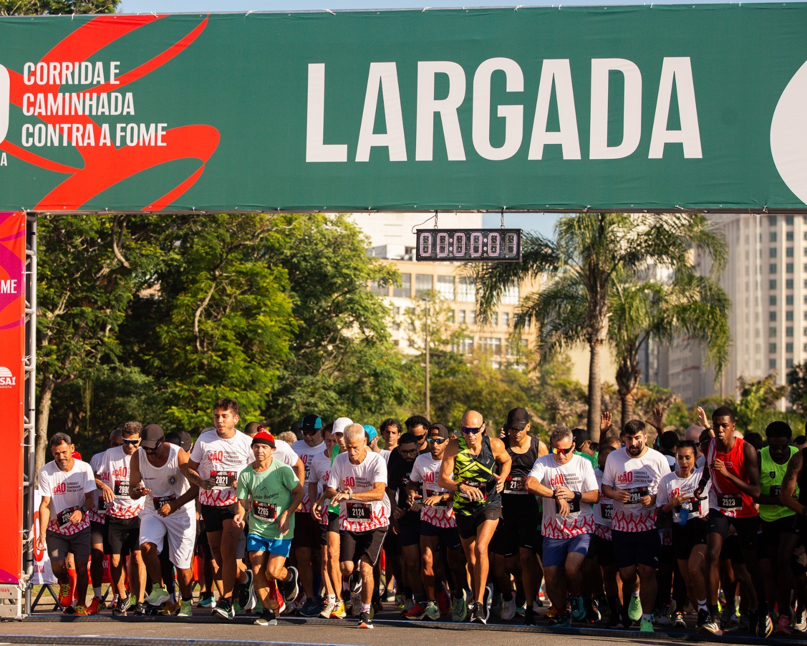 Corredores de diversas idades e etnias, vestindo camisetas brancas e coloridas, se preparam para a largada de uma corrida de rua sob um banner verde com os dizeres CORRIDA E CAMINHADA CONTRA A FOME e LARGADA, com um cronômetro digital marcando 00:00:00 acima deles. O cenário inclui árvores e prédios ao fundo, sob luz solar