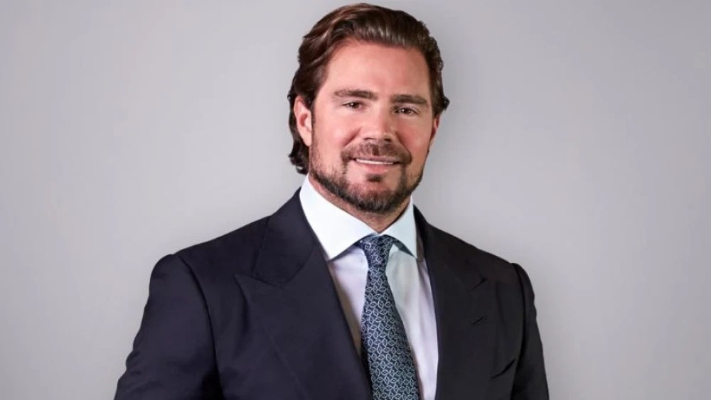 Homem branco de meia-idade, cabelo castanho penteado para trás, barba e bigode bem aparados, sorrindo levemente. Veste terno azul-marinho, camisa social branca e gravata azul com estampa geométrica, em fundo cinza claro