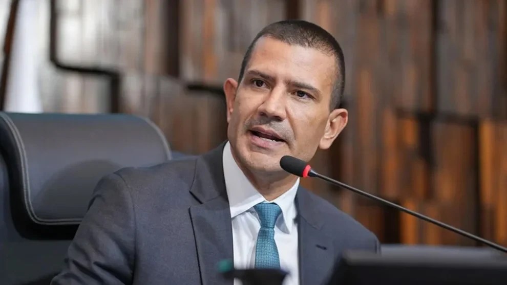 Homem de pele clara, cabelo curto e escuro, vestindo terno cinza e gravata azul-petróleo, fala em um microfone preto com detalhe vermelho, em um ambiente formal com painéis de madeira ao fundo