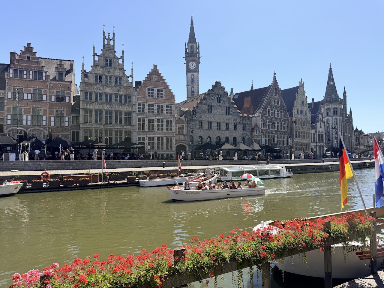 Cenário urbano com edifícios históricos de telhados pontiagudos e uma torre de relógio alta, margeando um rio onde barcos de passeio com pessoas navegam. Há bandeiras da Alemanha e Holanda à direita, e floreiras com flores vermelhas em primeiro plano, sob céu azul claro