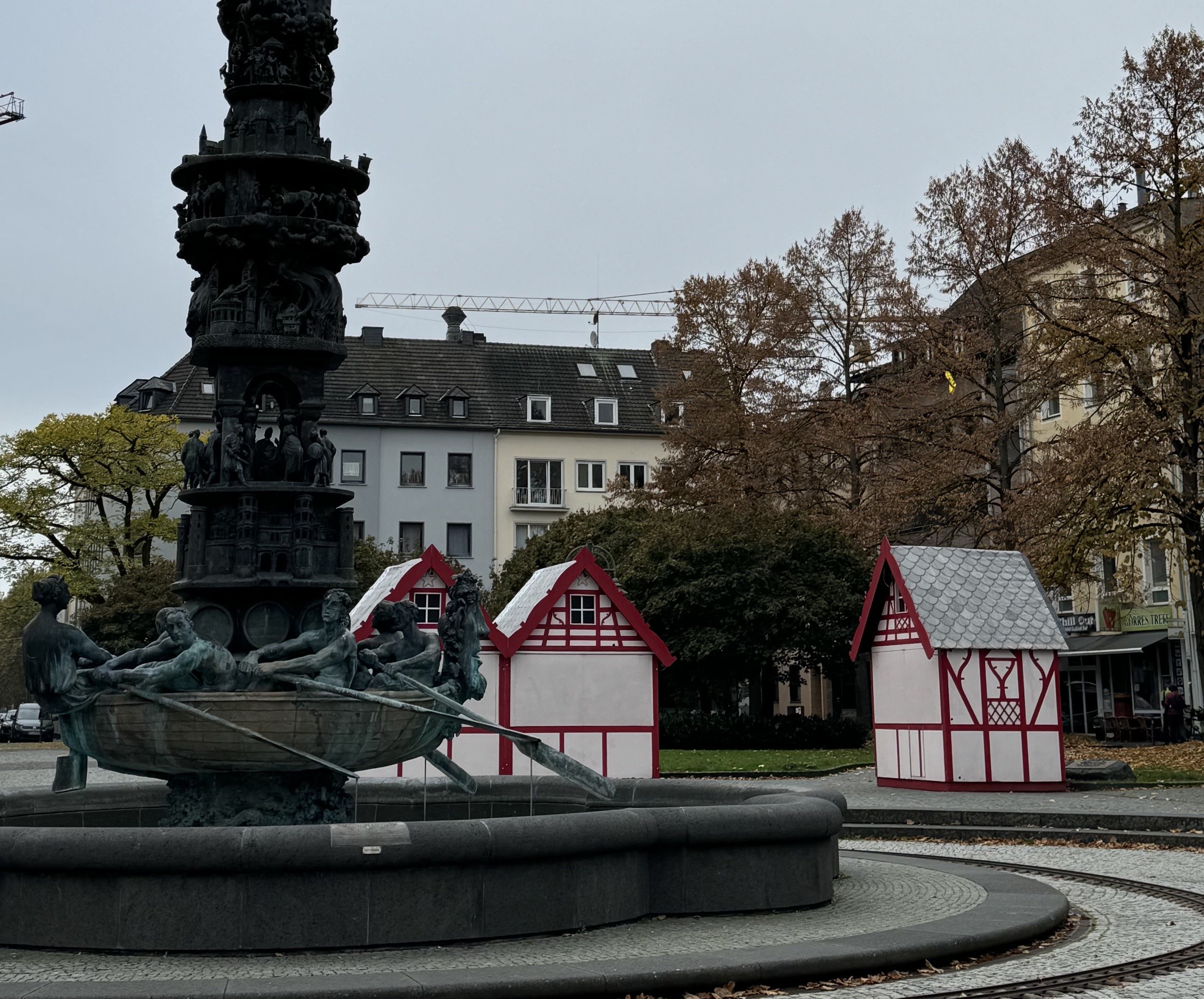 Fonte escura com esculturas de pessoas em um barco, ao lado de duas pequenas casas brancas com telhados vermelhos e detalhes em madeira, em uma praça com árvores de folhas secas e prédios ao fundo, sob céu nublado