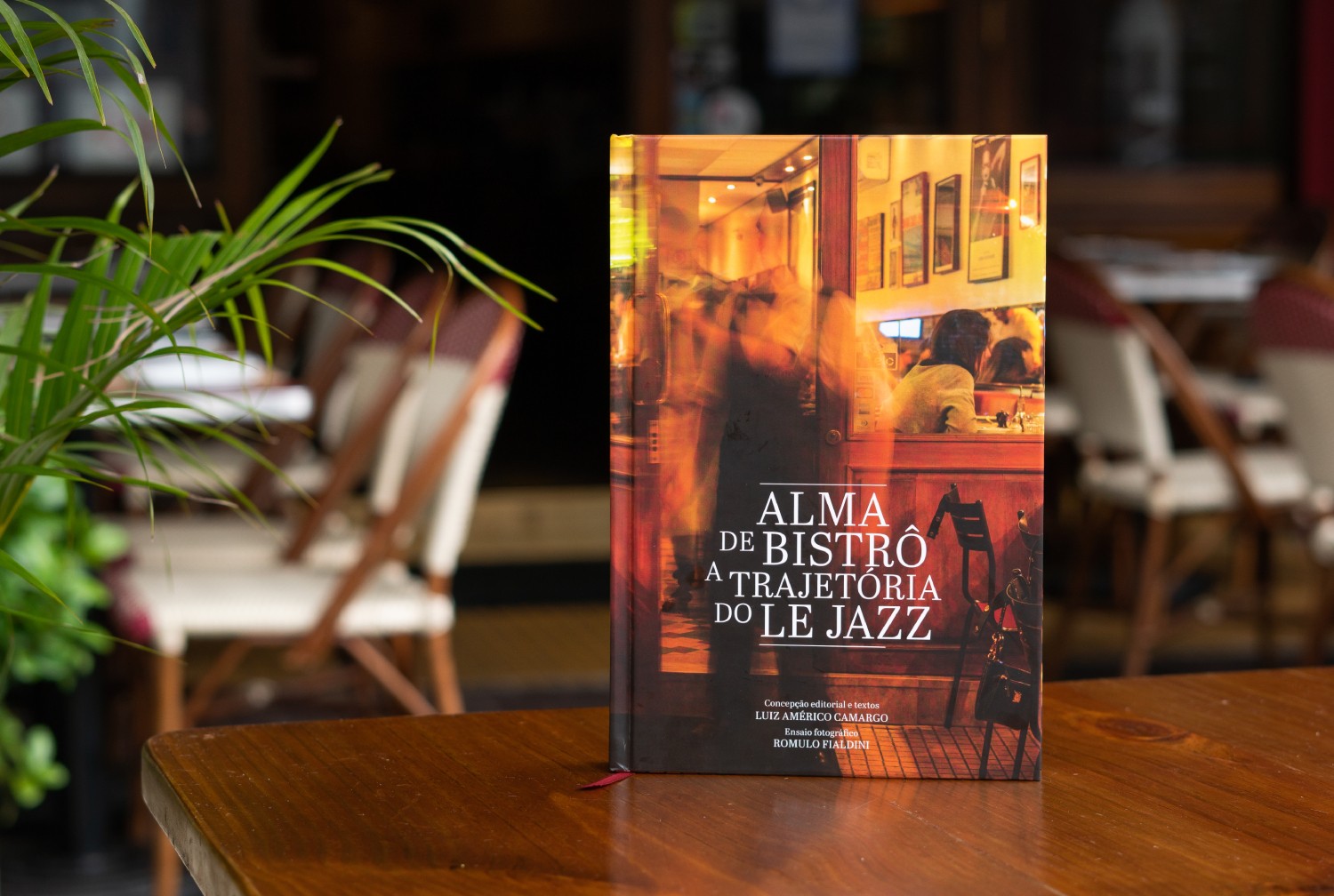 Livro Alma de Bistrô: A Trajetória do Le Jazz em capa dura, com imagem de um bistrô iluminado e pessoas borradas, sobre uma mesa de madeira escura. Folhas verdes de uma planta à esquerda, e cadeiras de madeira desfocadas ao fundo.