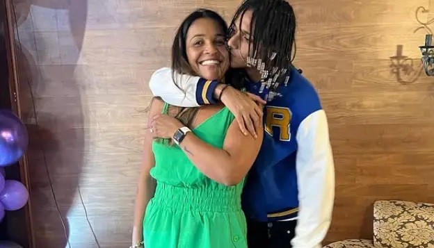 Mulher sorridente de vestido verde e relógio branco, abraçada por um homem de dreads e jaqueta azul e branca, que a beija na bochecha, em um ambiente com parede de madeira e balões roxos