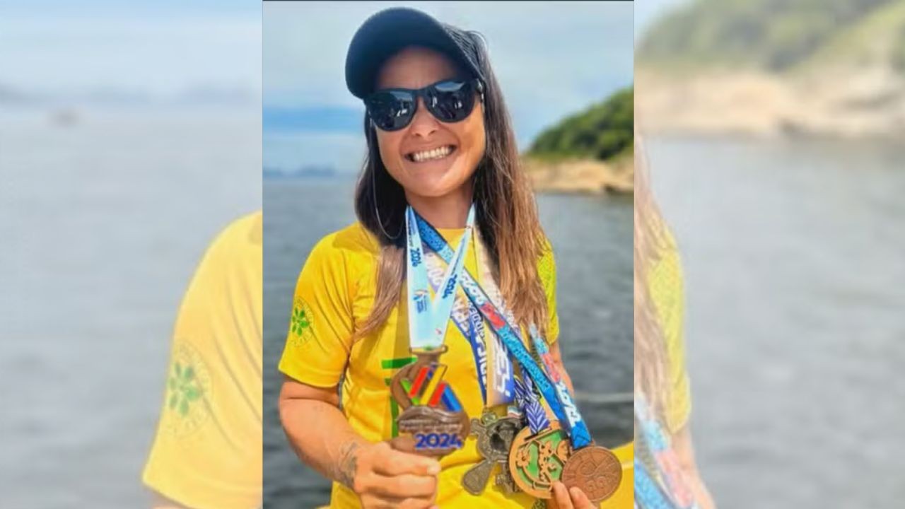 Mulher sorridente, de boné preto e óculos escuros, usando camiseta amarela e exibindo várias medalhas coloridas no pescoço e na mão, com um lago e vegetação ao fundo