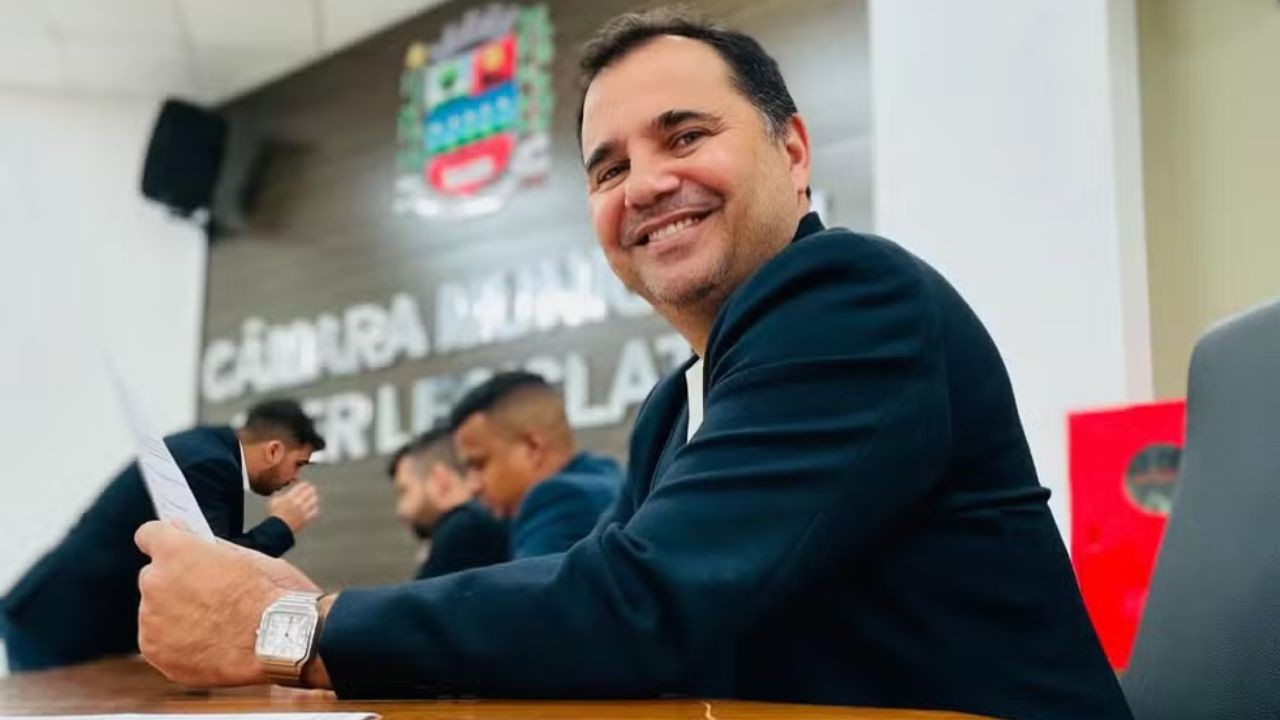 Homem de meia-idade, pele clara, cabelo escuro e sorriso largo, vestindo terno azul-marinho, sentado em uma mesa de madeira, segurando papéis. Ao fundo, outros homens e o letreiro CÂMARA MUNICIPAL LEGISLATIVA com um brasão