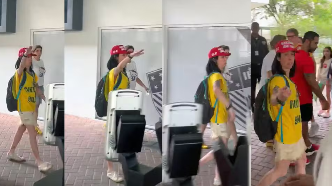Mulher de cabelo escuro, boné vermelho e camiseta amarela com mochila preta, caminhando em ambiente interno, gesticulando com a mão direita estendida, enquanto outras pessoas transitam ao fundo