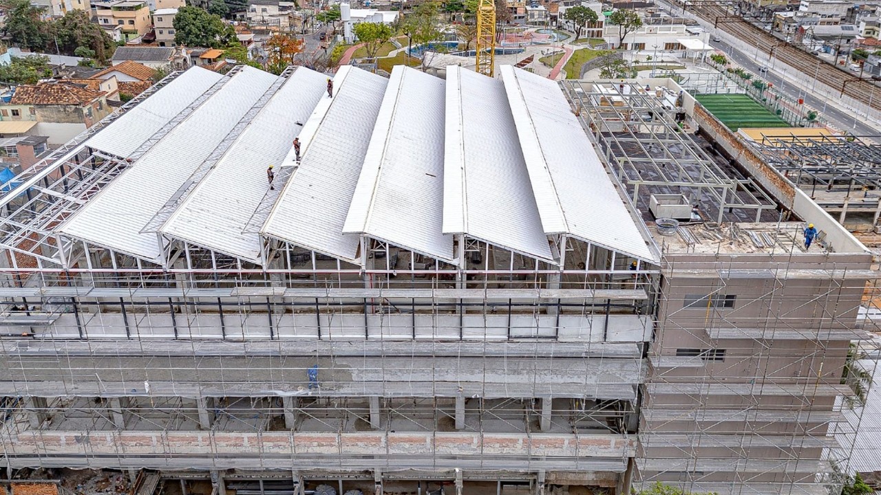Vista aérea de um edifício em construção com telhado branco em formato de serra, andaimes ao redor e trabalhadores no topo. Ao fundo, casas e uma área verde com piscina.