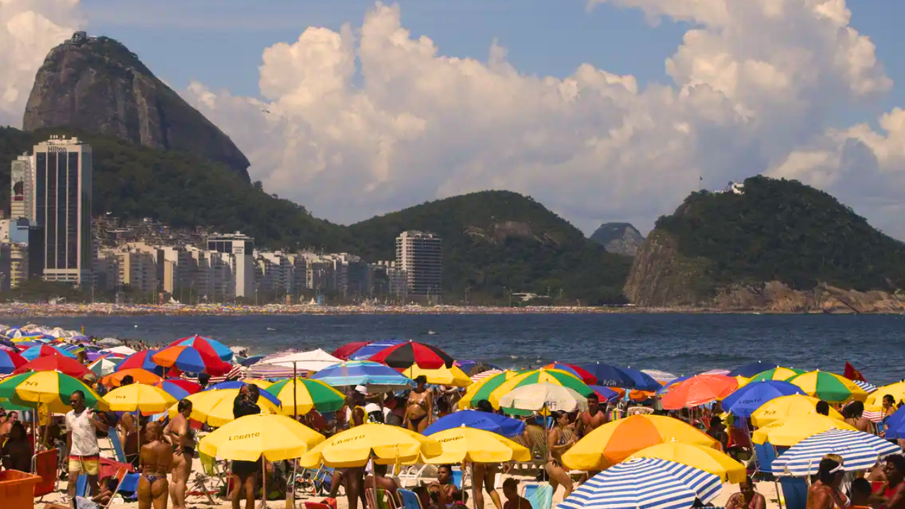 Praia lotada com guarda-sóis coloridos e pessoas, o mar azul ao fundo. Montanhas verdes e prédios altos da cidade completam a paisagem sob um céu com nuvens brancas