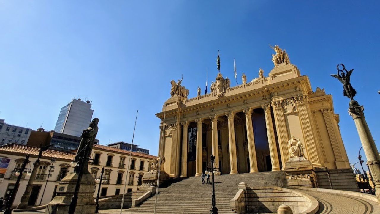 Fachada imponente do Palácio da Liberdade em Belo Horizonte, com colunas clássicas, estátuas no topo e escadaria ampla, sob céu azul claro. Edifícios modernos e antigos ao redor, com uma estátua escura em primeiro plano à esquerda e outra em um pilar alto à direita