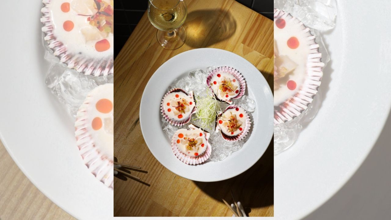 Quatro vieiras frescas em conchas brancas e roxas, com pontos vermelhos e cebolinha, servidas sobre gelo em um prato branco, ao lado de uma taça de vinho branco em uma mesa de madeira