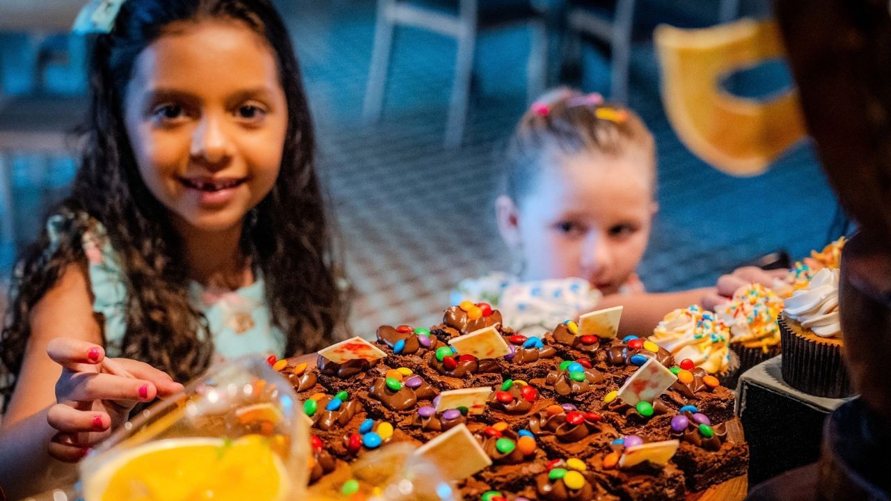 Duas meninas, uma com cabelo cacheado e outra loira, observam uma mesa cheia de doces, incluindo um bolo de chocolate com confeitos coloridos e cupcakes. A menina de cabelo cacheado sorri para a câmera, enquanto a outra olha curiosa para os doces.