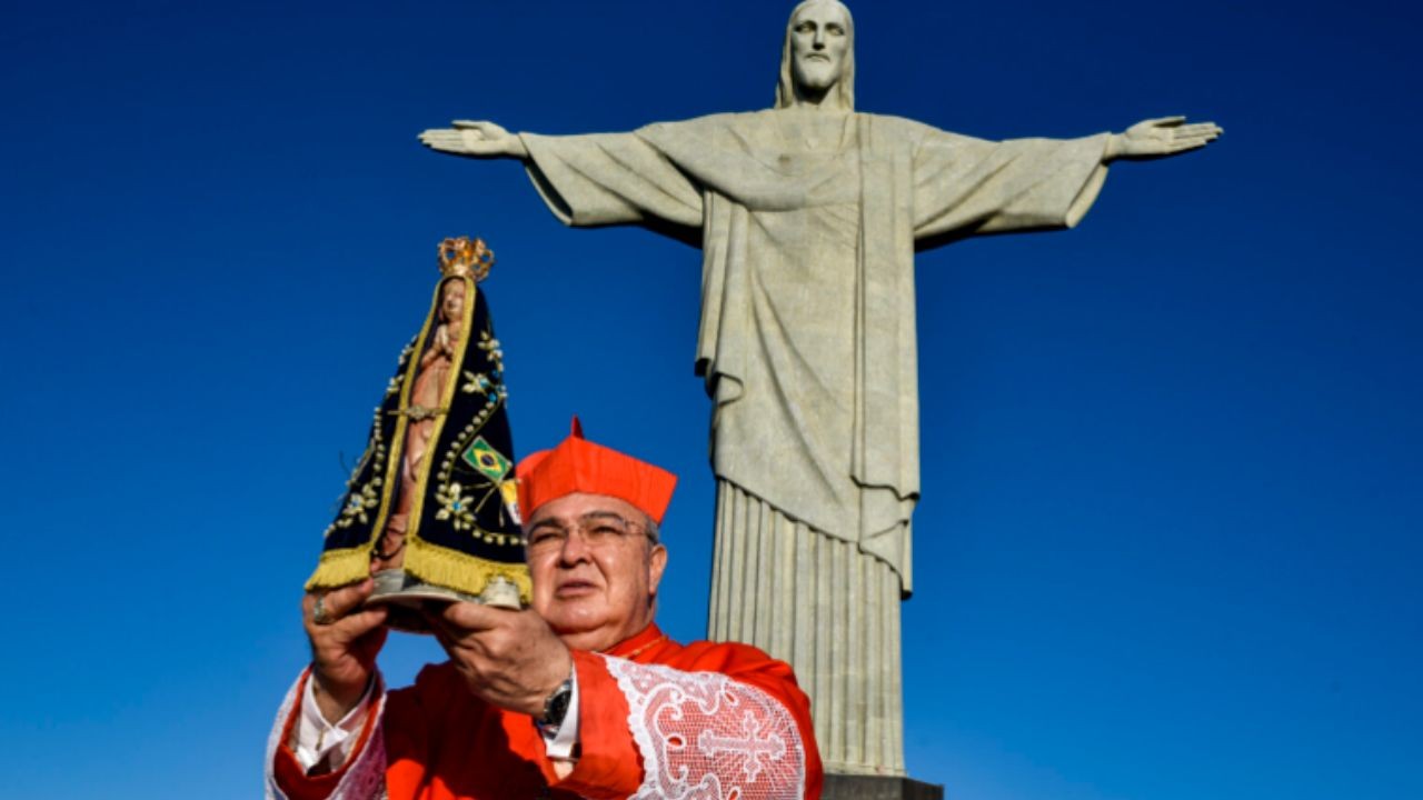 Cardeal brasileiro de batina vermelha e barrete, segurando uma imagem de Nossa Senhora Aparecida com manto decorado com a bandeira do Brasil, em frente ao Cristo Redentor sob céu azul