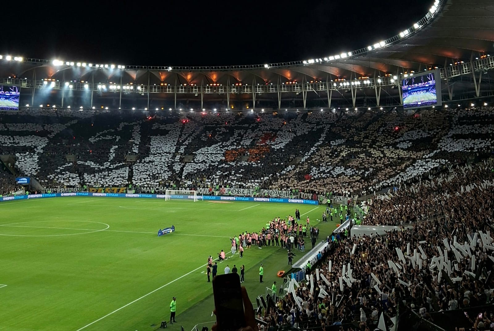 Estádio de futebol lotado à noite, com torcedores nas arquibancadas formando um mosaico preto e branco. O campo verde iluminado tem jogadores e equipe técnica na lateral. Telões exibem imagens no alto.