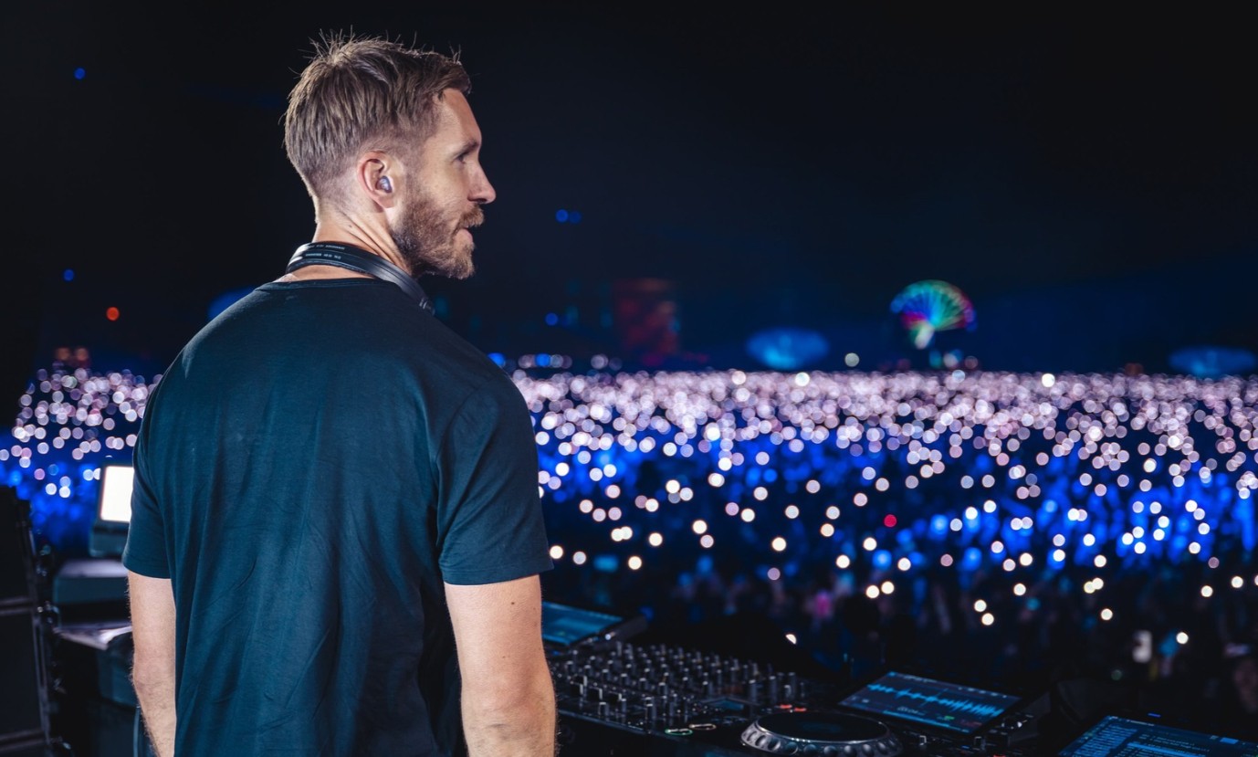 DJ Calvin Harris de costas, usando camiseta azul-marinho e fones de ouvido no pescoço, olhando para uma multidão iluminada por luzes azuis e brancas em um show noturno